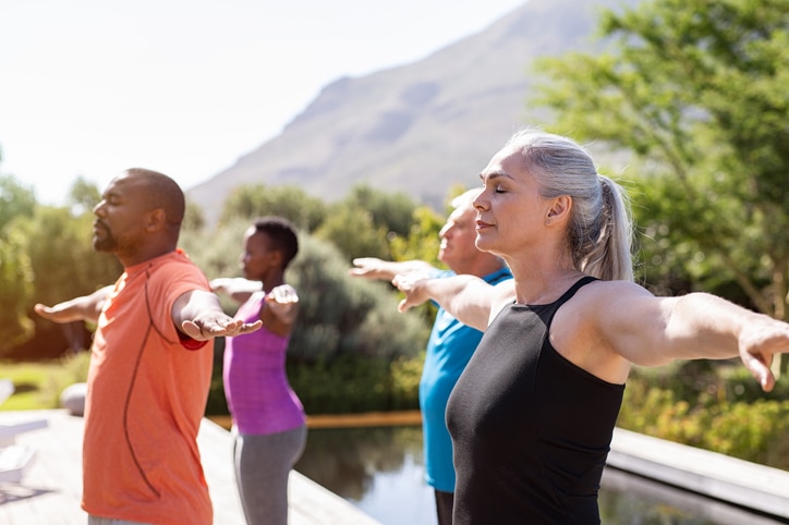 A group of older adults doing the same yoga pose in front of a small pool and mountainous landscape