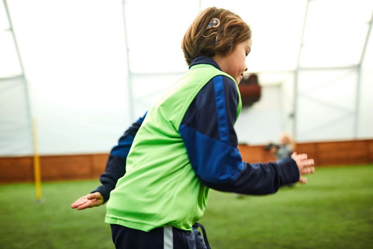 A child with a cochlear implant running across an indoor practice field